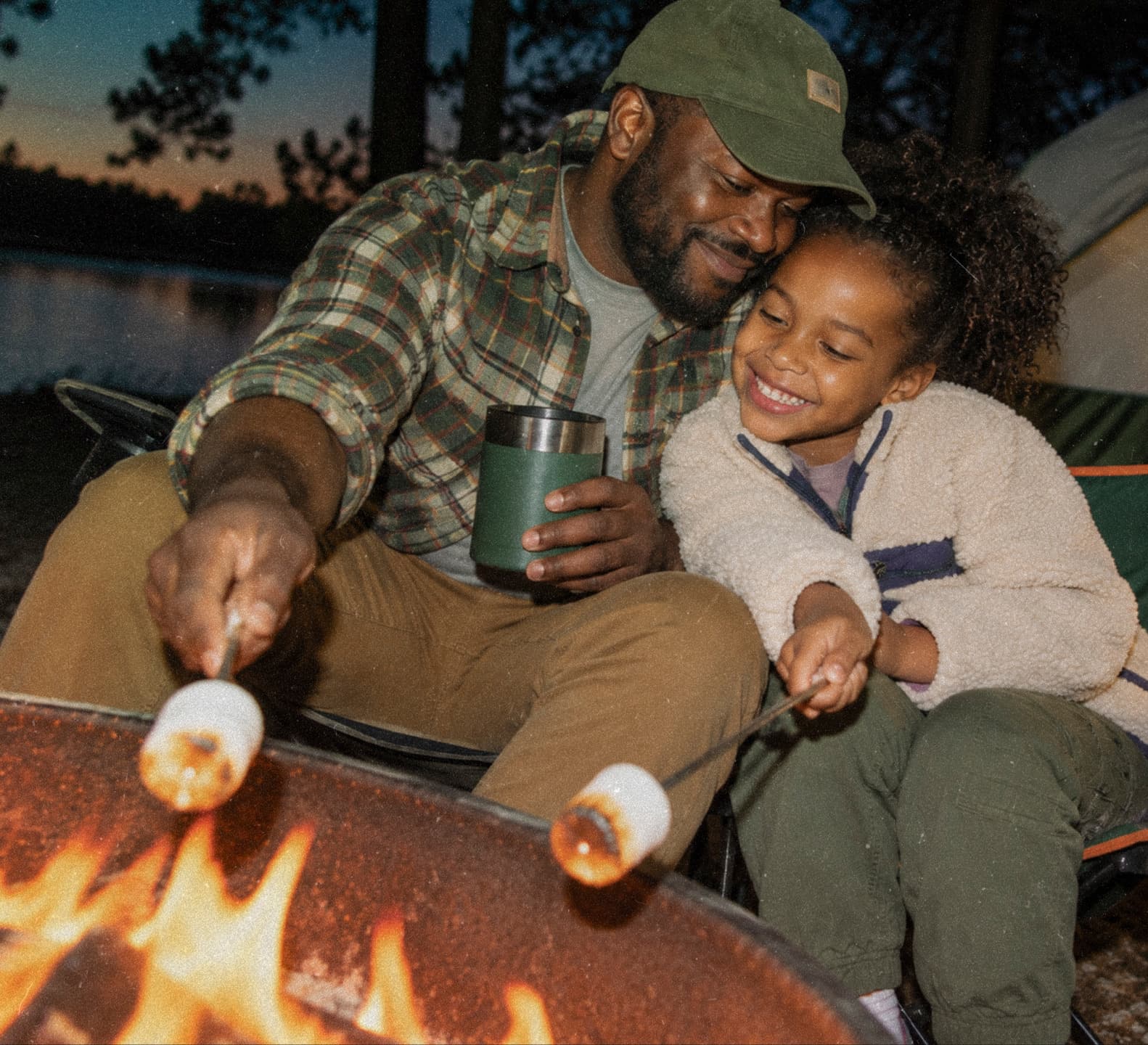 A father and daughter laughing as they toast marshmallows over a campfire.