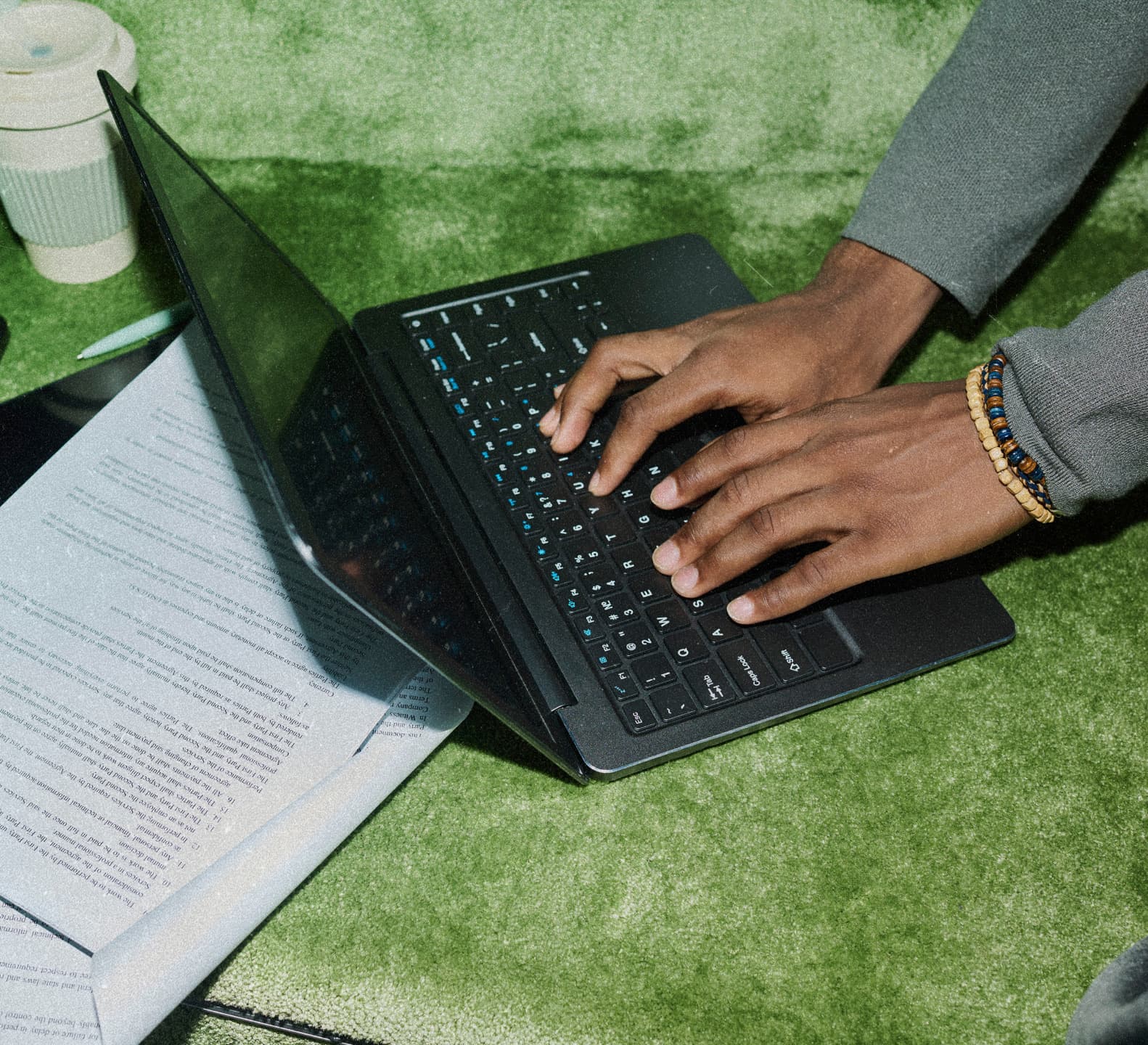 Hands typing on a laptop on a green rug beside printed pages, a tablet, and a coffee cup.