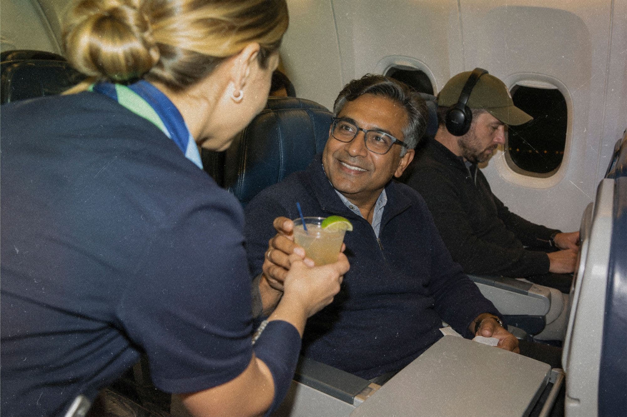 A flight attendant hands a smiling passenger a drink with a lime wedge.