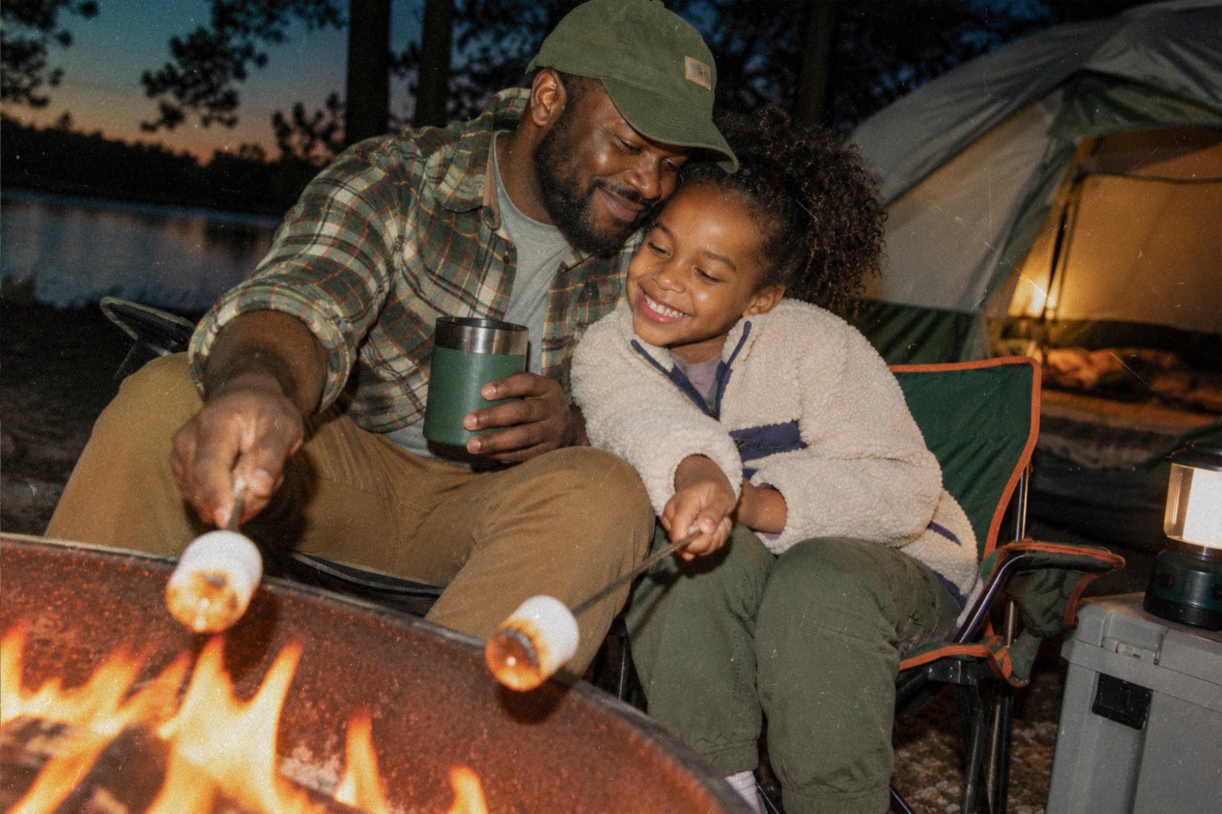 A father and daughter laughing as they toast marshmallows over a campfire.