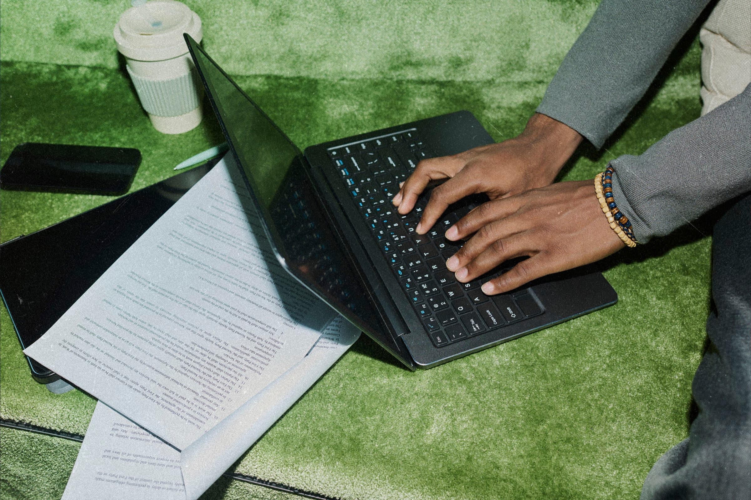 Hands typing on a laptop on a green rug beside printed pages, a tablet, and a coffee cup.
